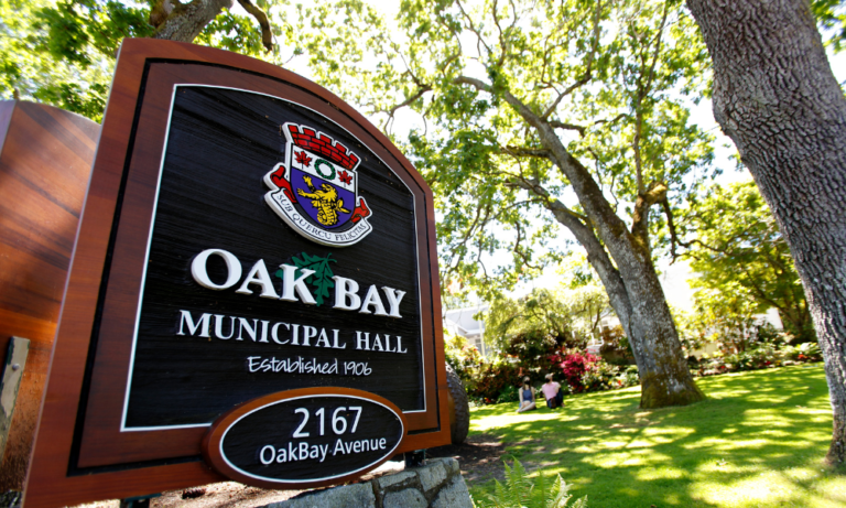 A sign for Oak Bay Municipal Hall with the Oak Bay crest is in the foreground with large trees with green leaves, and grass in the background including Oak Bay's Municipal Hall.