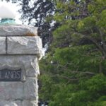 A stone column with the work "Uplands" on a plaque, with a green street light pole in the background bordered by trees.