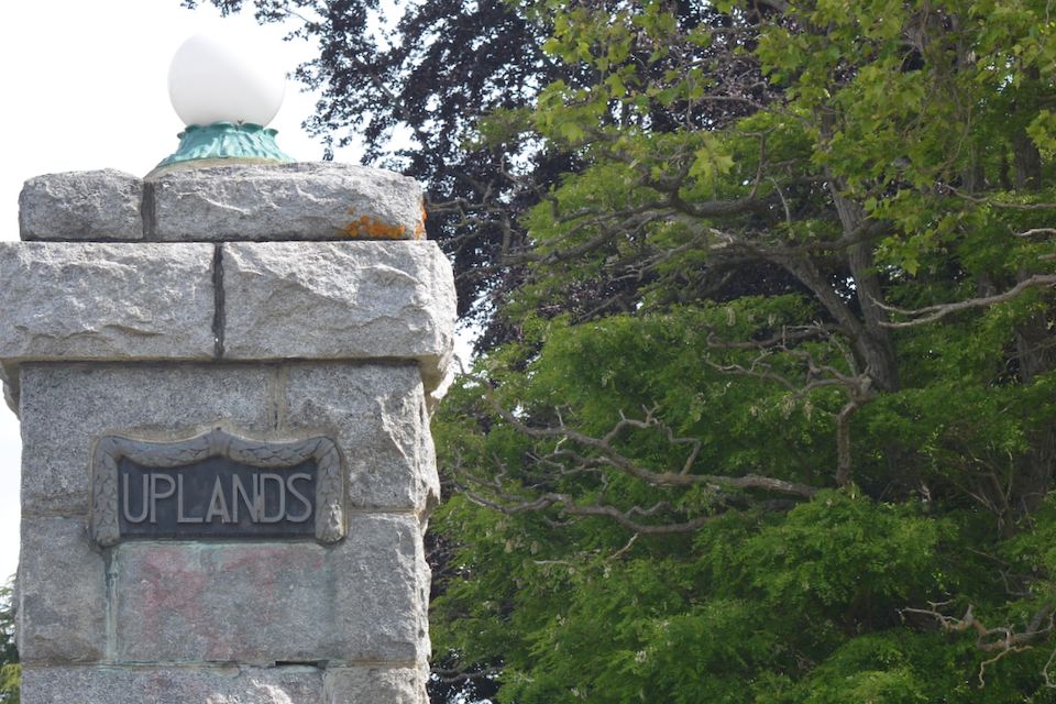 A stone column with the work "Uplands" on a plaque, with a green street light pole in the background bordered by trees.