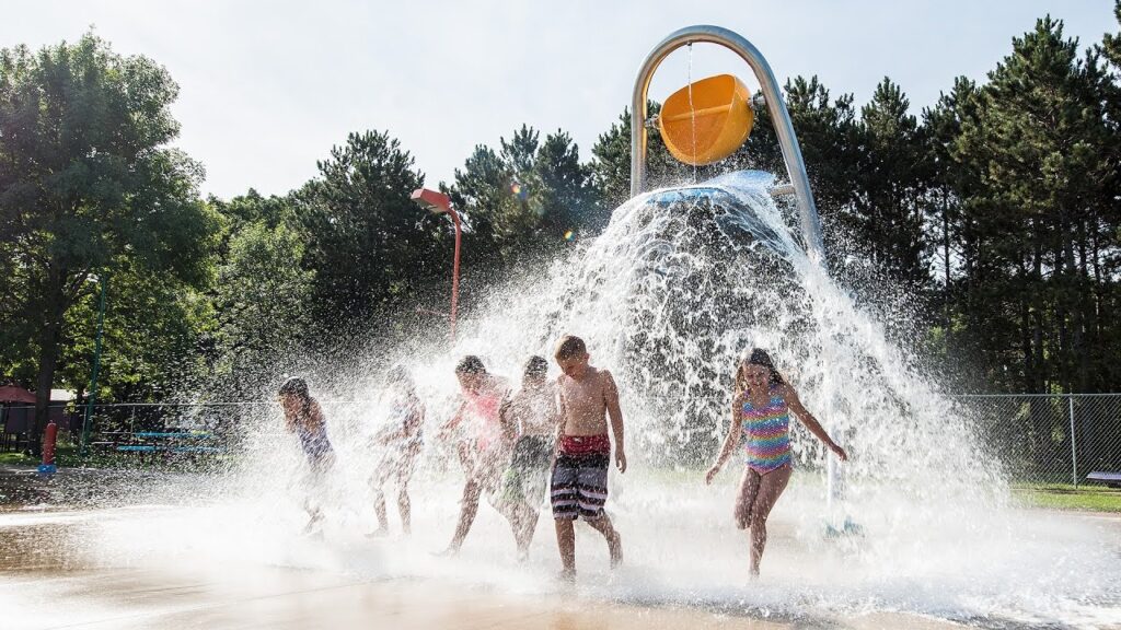 Children run through a splash park with a giant bucket on poles dumping water on them.