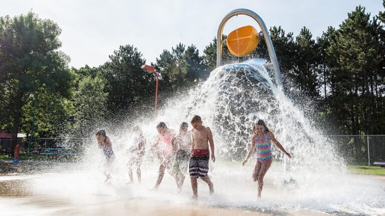Children run through a splash park with a giant bucket on poles dumping water on them.