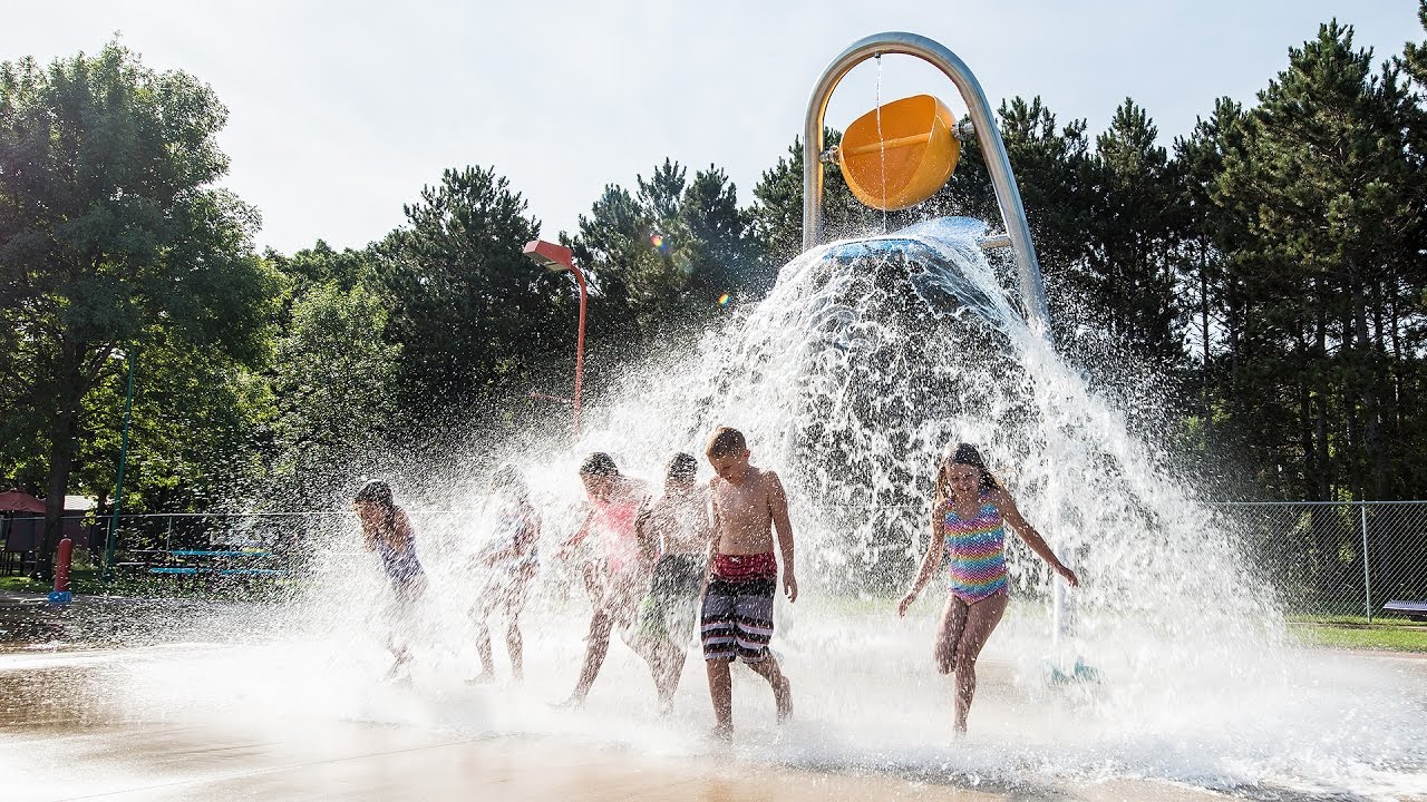 Children run through a splash park with a giant bucket on poles dumping water on them.