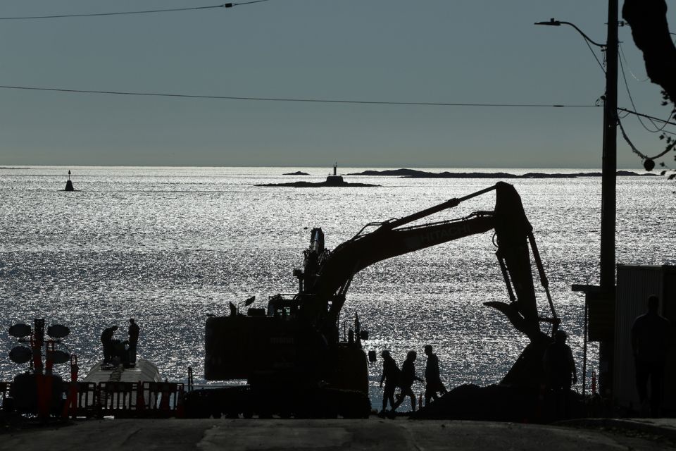 A large backhoe is parked on a beach with the ocean in the background and people walking along a sidewalk next to the beach.