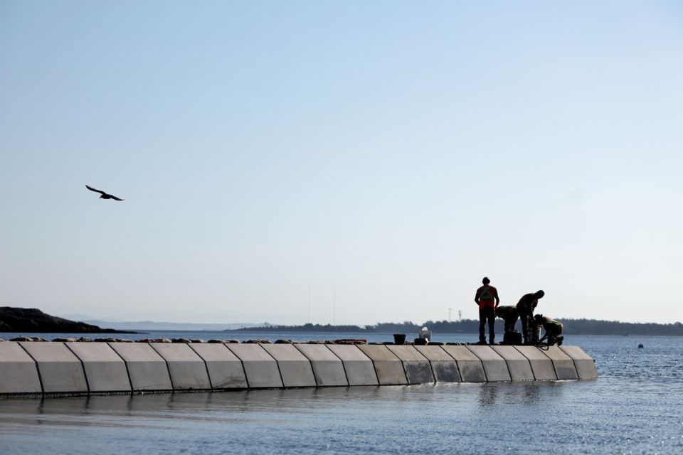 A concrete covering of a pipe extends out into the ocean, with crews standing on it and inspecting it. Blue sky and sun in the background with a bird flying above.